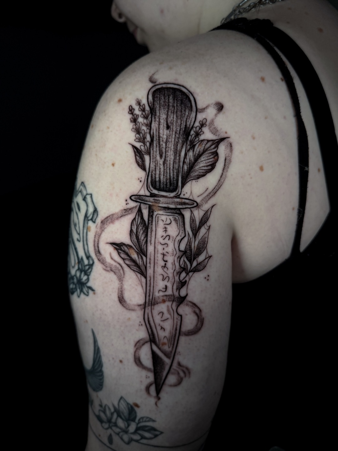 Zwart-grijze tattoo van een dolk met bladeren en een lint op een bovenarm.
