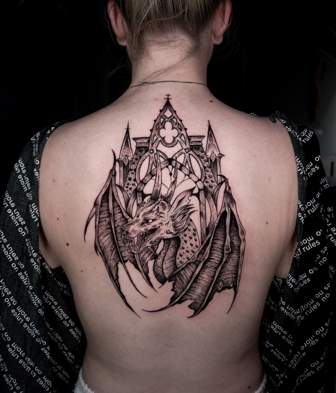Zwart-grijze tattoo van een draak voor een gotische kathedraalboog op een bovenrug.