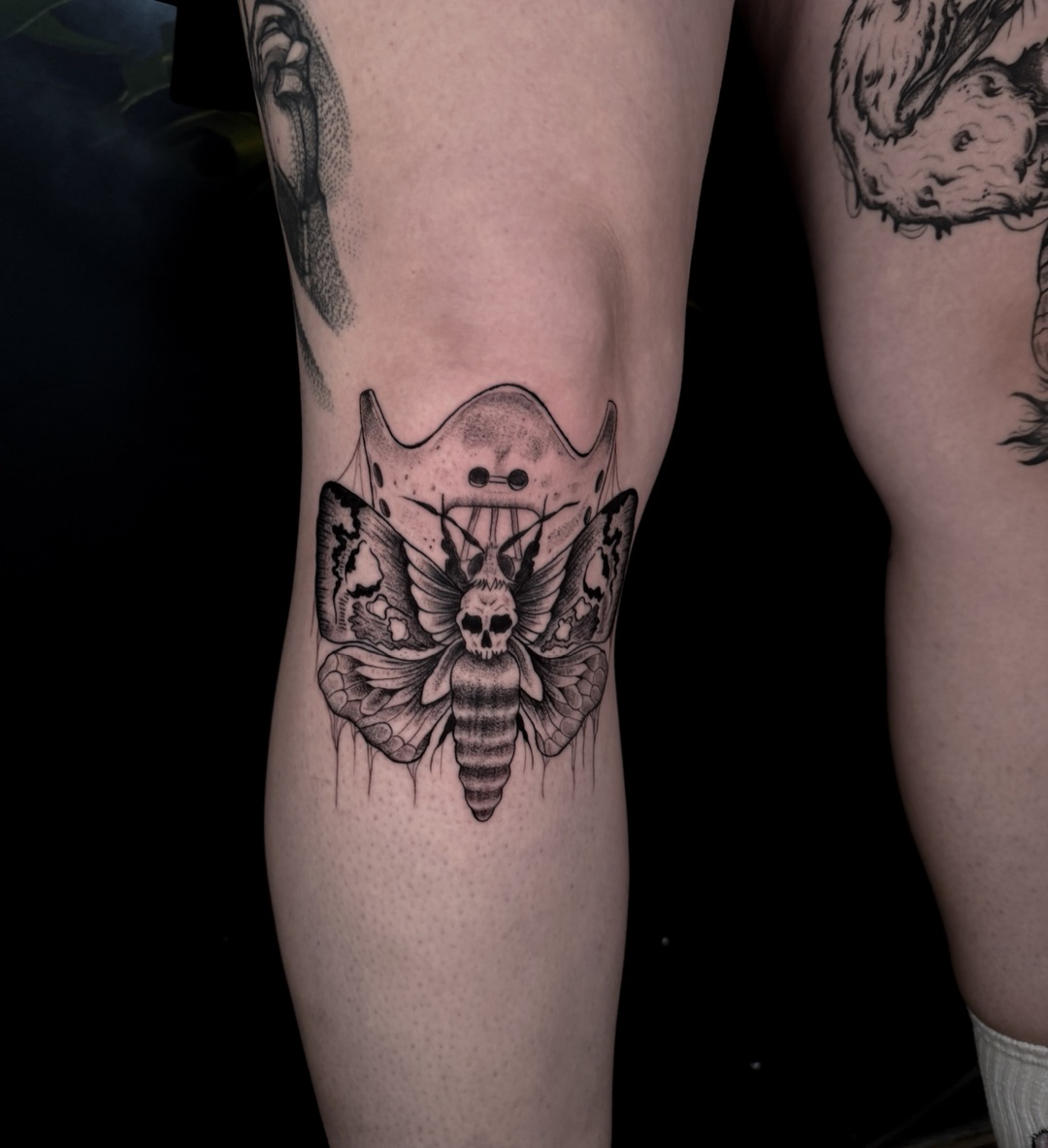 Zwart-grijze tattoo van een doodshoofdvlinder met Hannibal-masker op de achterkant van een knie.
