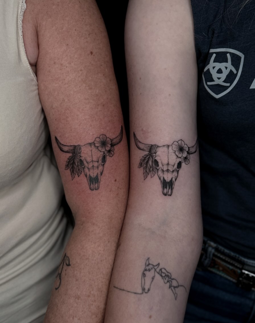 Twee onderarmen naast elkaar, elk met een fijnlijnige koeienschedel met bloemen en veren.